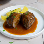 Rinderroulade auf Teller mit Sauce und Kartoffelspalten, garniert mit Petersilie.