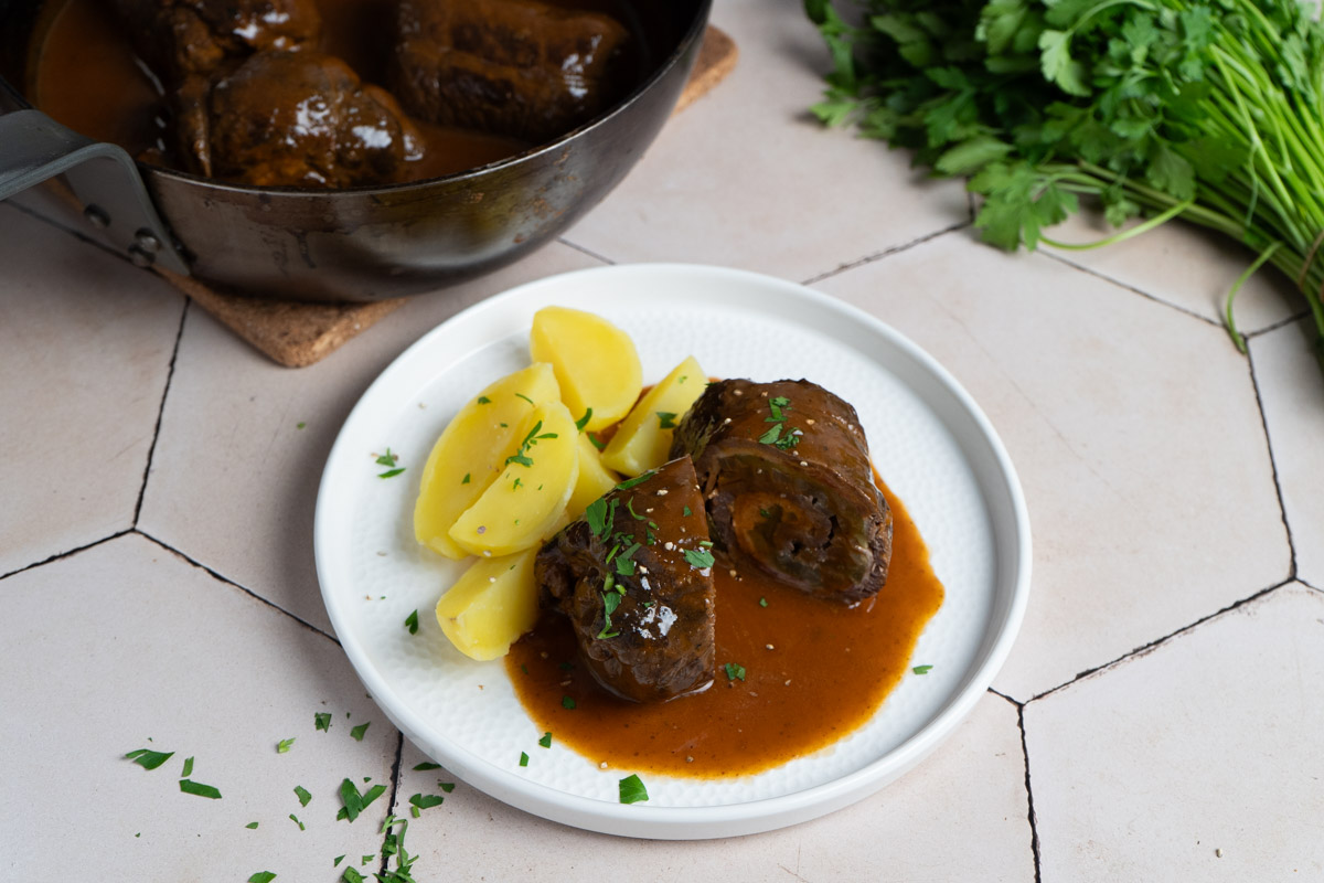 Roulade mit Soße und Kartoffeln auf Teller, daneben Topf mit Rouladen und frische Petersilie auf hellem Untergrund.