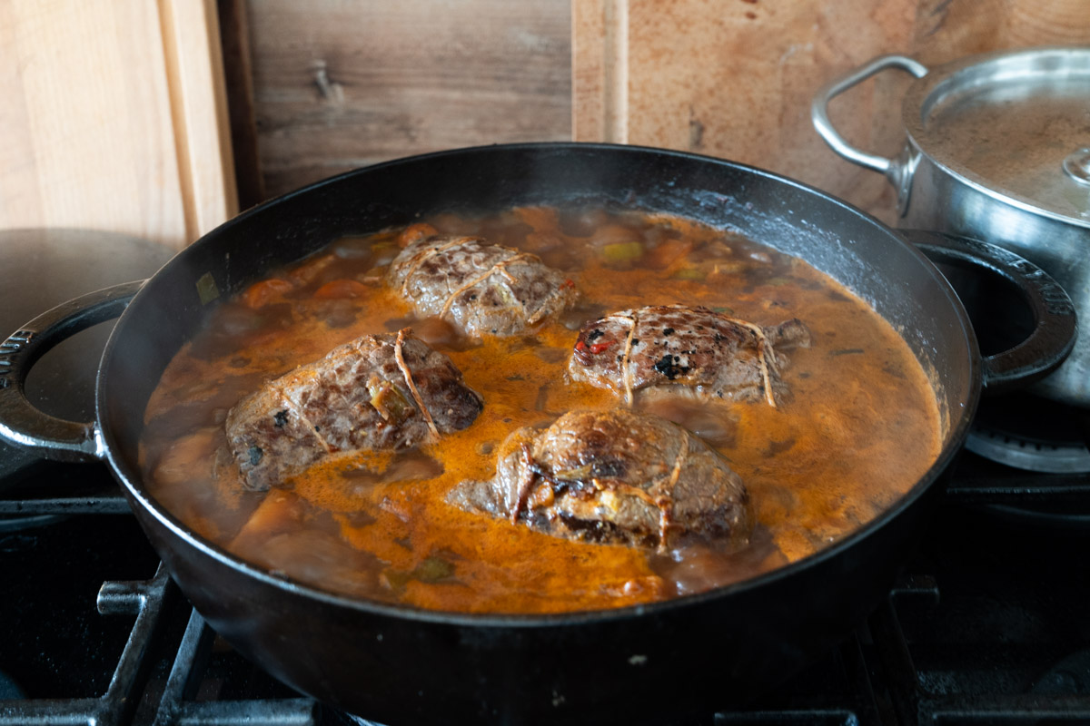 Große Pfanne auf Herd mit vier gefüllten Rinderrouladen in brauner Soße, die köcheln.