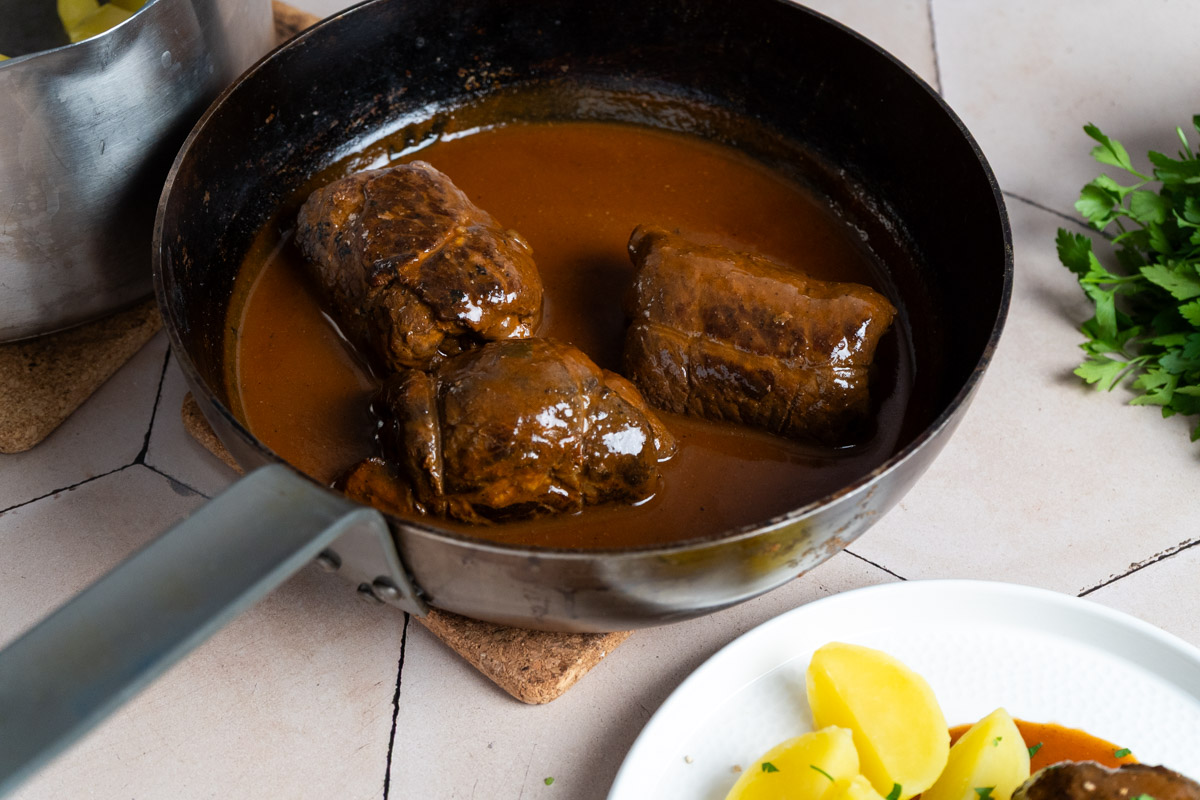 Pfanne mit drei braunen Rouladen in Soße, daneben gekochte Kartoffeln auf einem weißen Teller.