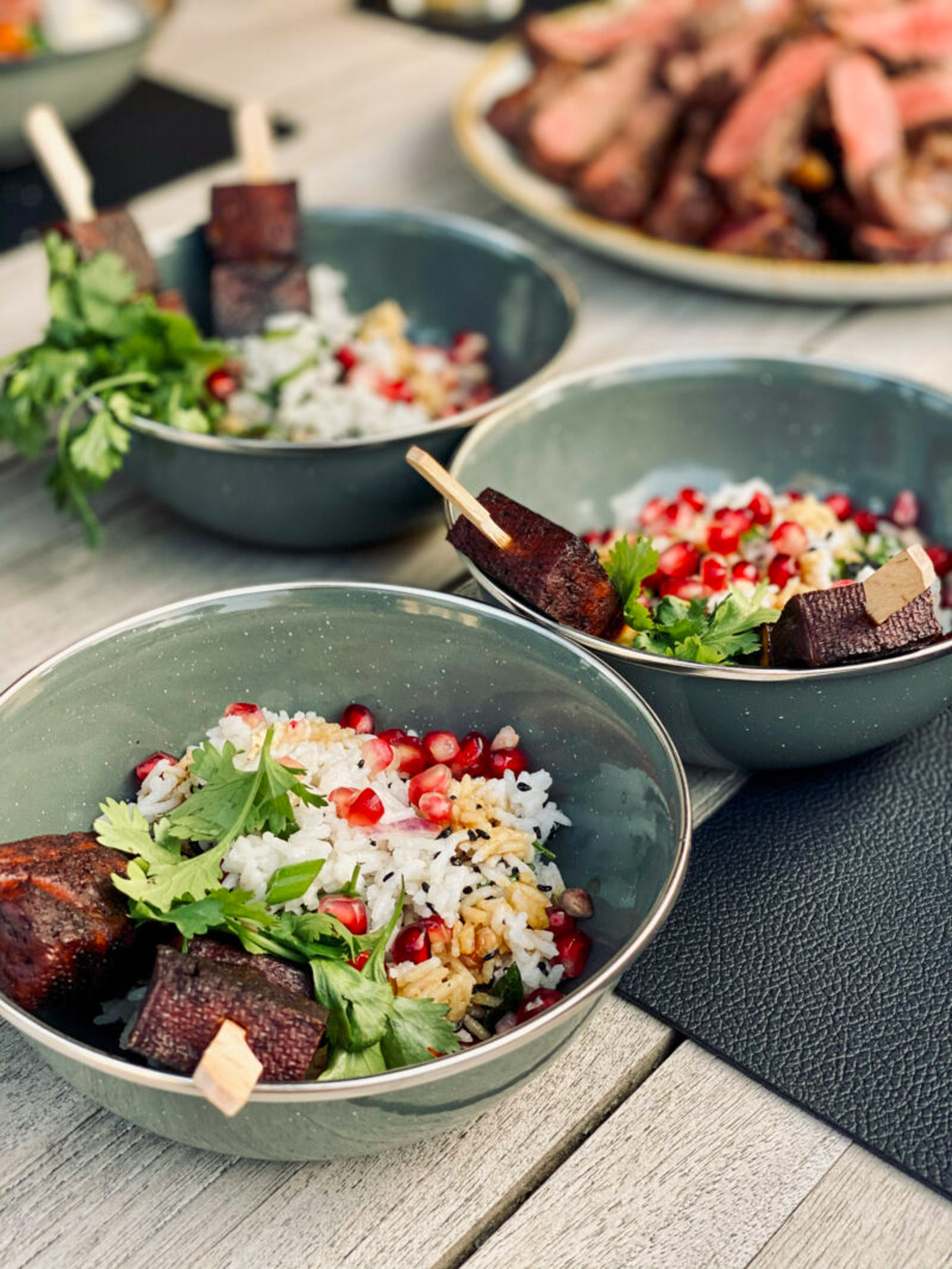 Teriyaki-Tofu mit Kokos-Basmatireis_01 Drei Schalen mit Reis, Granatapfel, Kräutern und gegrillten Fleischstücken auf Holzunterlage. Im Hintergrund weiteres Fleisch.