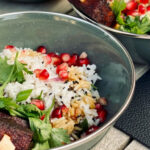 Zwei Schalen mit Reis, Granatapfelkernen, frischen Kräutern und gebratenen Würfeln auf Holz- und Lederunterlage.