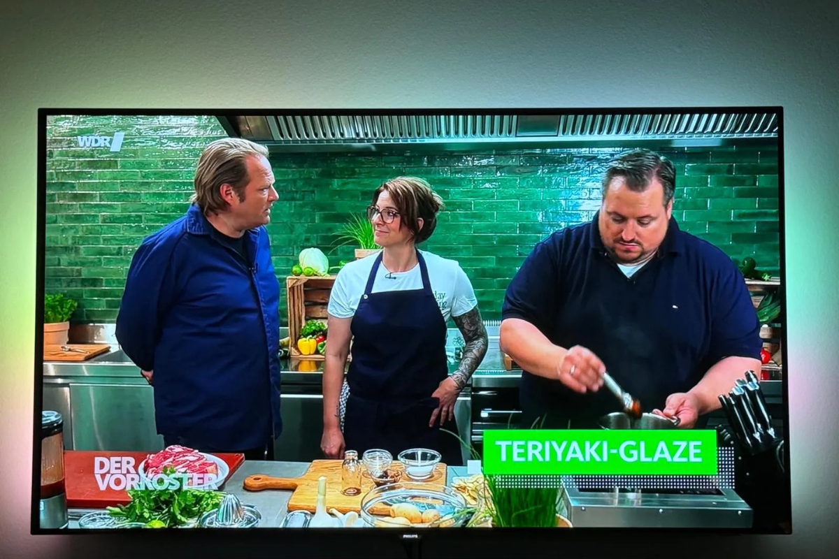 Drei Personen in einer TV-Kochshowküche, eine Person kocht, während die anderen zwei zusehen. Text: „TERIYAKI-GLAZE“.