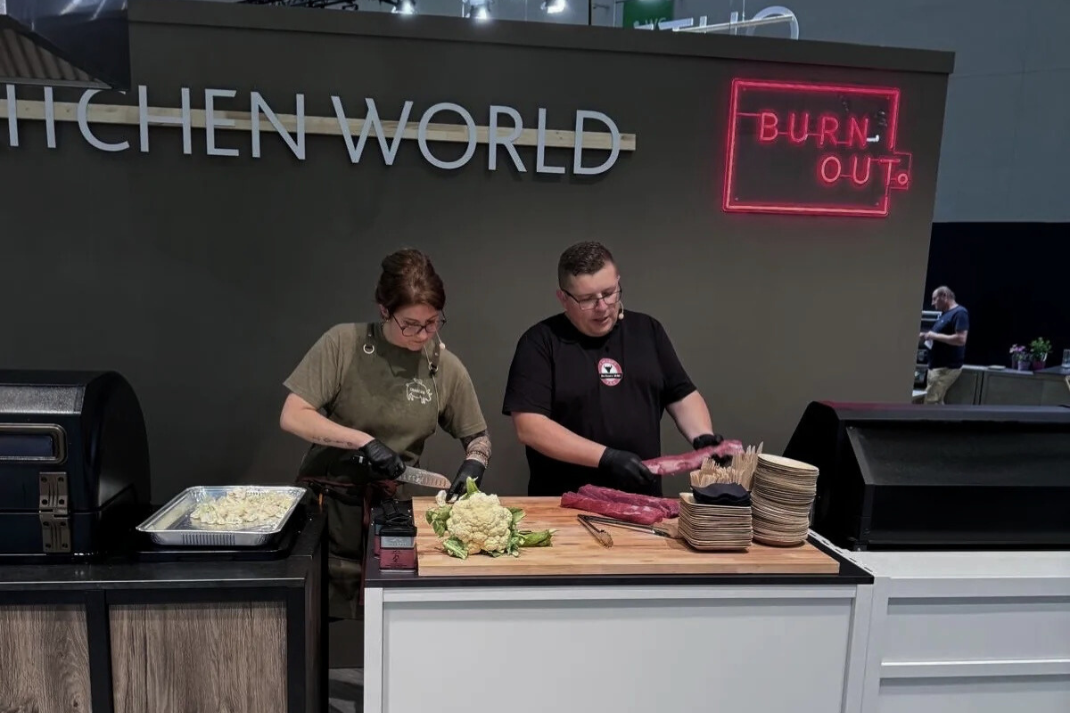 Zwei Personen bereiten auf einer Bühne mit Grill und Gemüse Speisen zu, erkennbar an Schürze und Kochwerkzeugen.