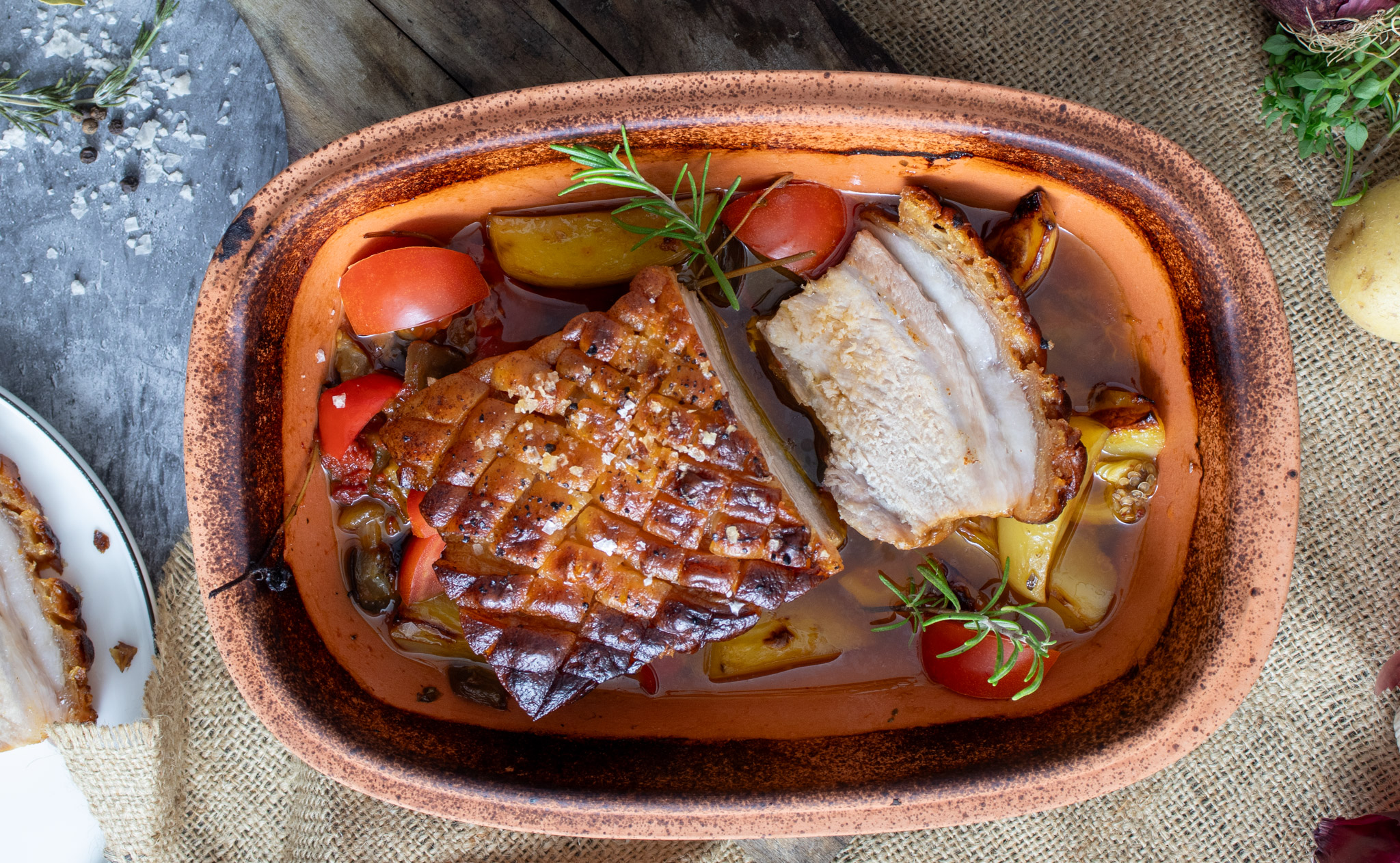 Gebackenes Schweinefleisch in einer Tonform, mit knuspriger Kruste, Tomaten und Rosmarin garniert.