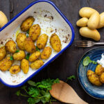 Gebackene Kartoffeln in Gewürzmischung im Ofenblech, serviert mit Kräuterdip auf einem Teller, umgeben von frischen Kräutern.