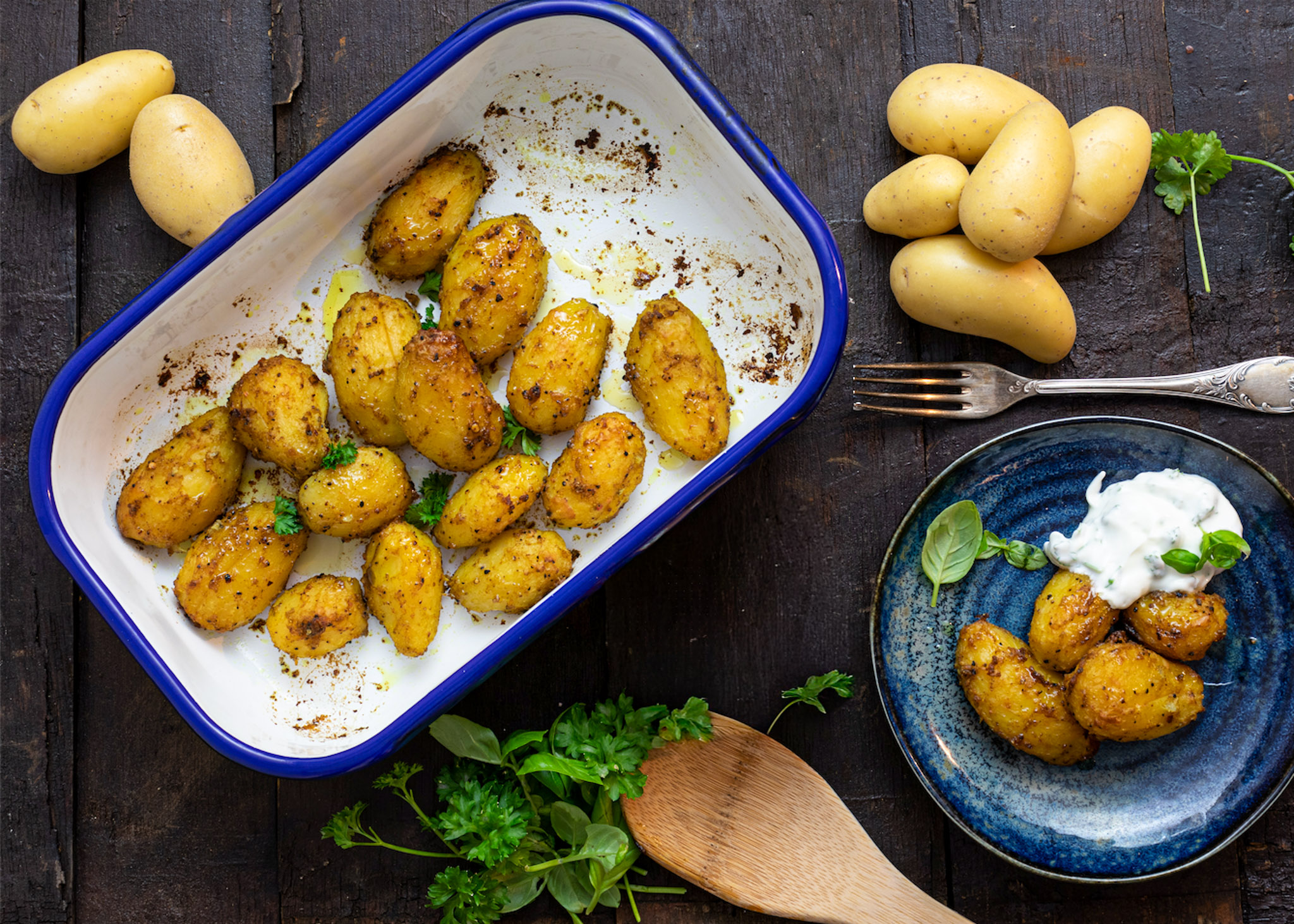 Gebackene Kartoffeln in Gewürzmischung im Ofenblech, serviert mit Kräuterdip auf einem Teller, umgeben von frischen Kräutern.