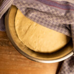 Teigschüssel mit aufgegangenem Hefeteig, teils mit einem lila Geschirrtuch abgedeckt, auf einem Holztisch.