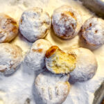 Acht mit Puderzucker bestäubte Mini-Quarkbällchen, eines mit einem Biss, in einer rechteckigen Metallform.