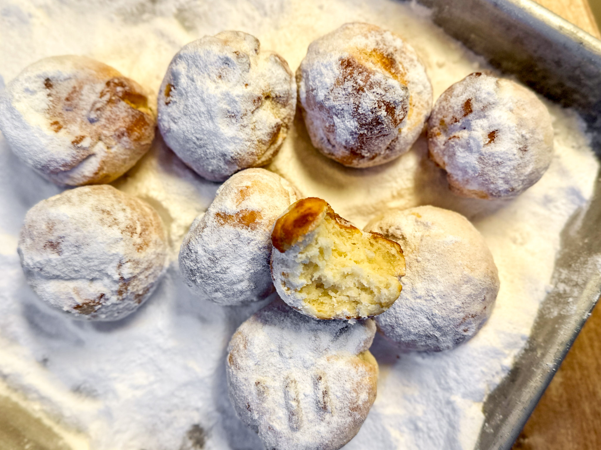 Quarkbällchen-aus-der-Heißluftfritteuse-01 Acht mit Puderzucker bestäubte Mini-Quarkbällchen, eines mit einem Biss, in einer rechteckigen Metallform.