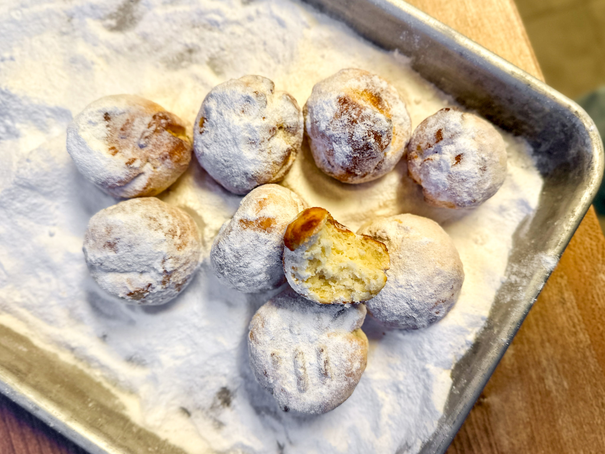 Acht mit Puderzucker bestäubte Gebäckbällchen auf einem Tablett, eins ist angeschnitten und zeigt die weiche Füllung.