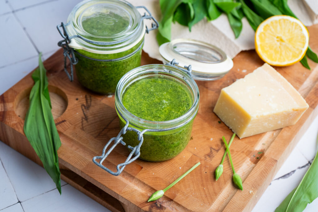 Zwei Gläser grünes Pesto, ein Stück Käse, Bärlauchblätter und eine halbe Zitrone auf einem Holzbrett.