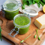Zwei Gläser grünes Pesto, ein Stück Käse, Bärlauchblätter und eine halbe Zitrone auf einem Holzbrett.