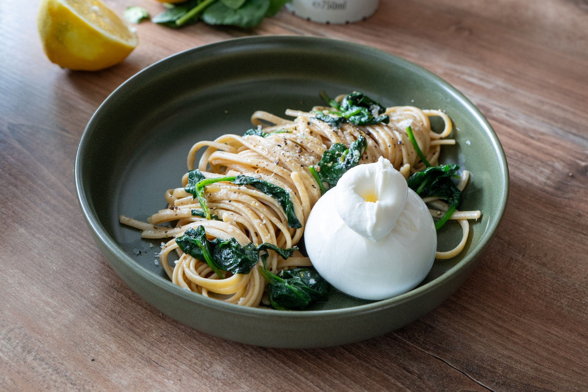 Nudeln mit Spinat und Pfeffer, serviert mit einer Kugel Burrata auf einem grünen Teller; Zitrone im Hintergrund.