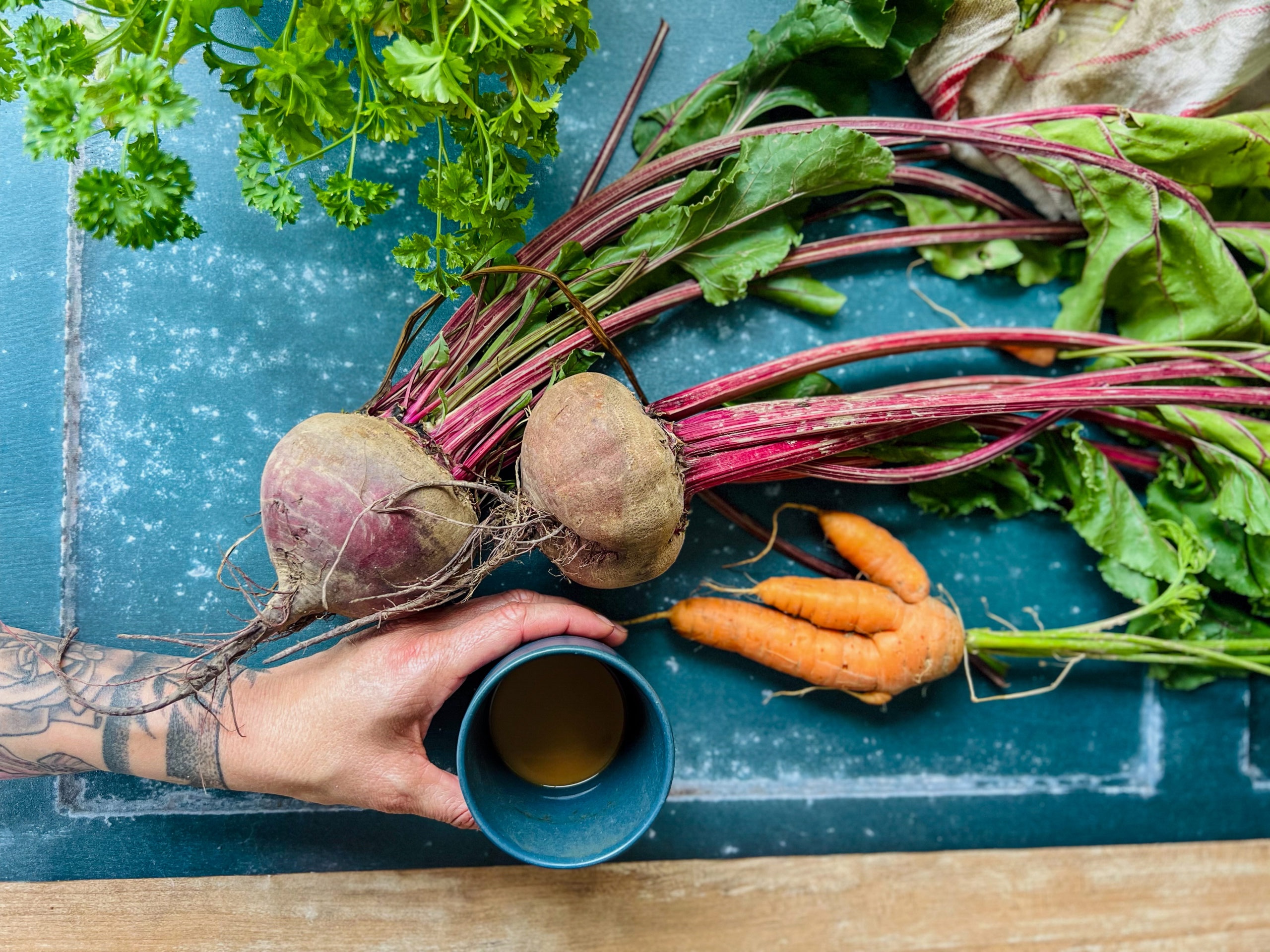 Rote_Bete_Risotto_04 Tattooierte Hand hält blaue Tasse neben frischem Gemüse, darunter Rote Bete, Karotten und Petersilie auf blauem Untergrund.