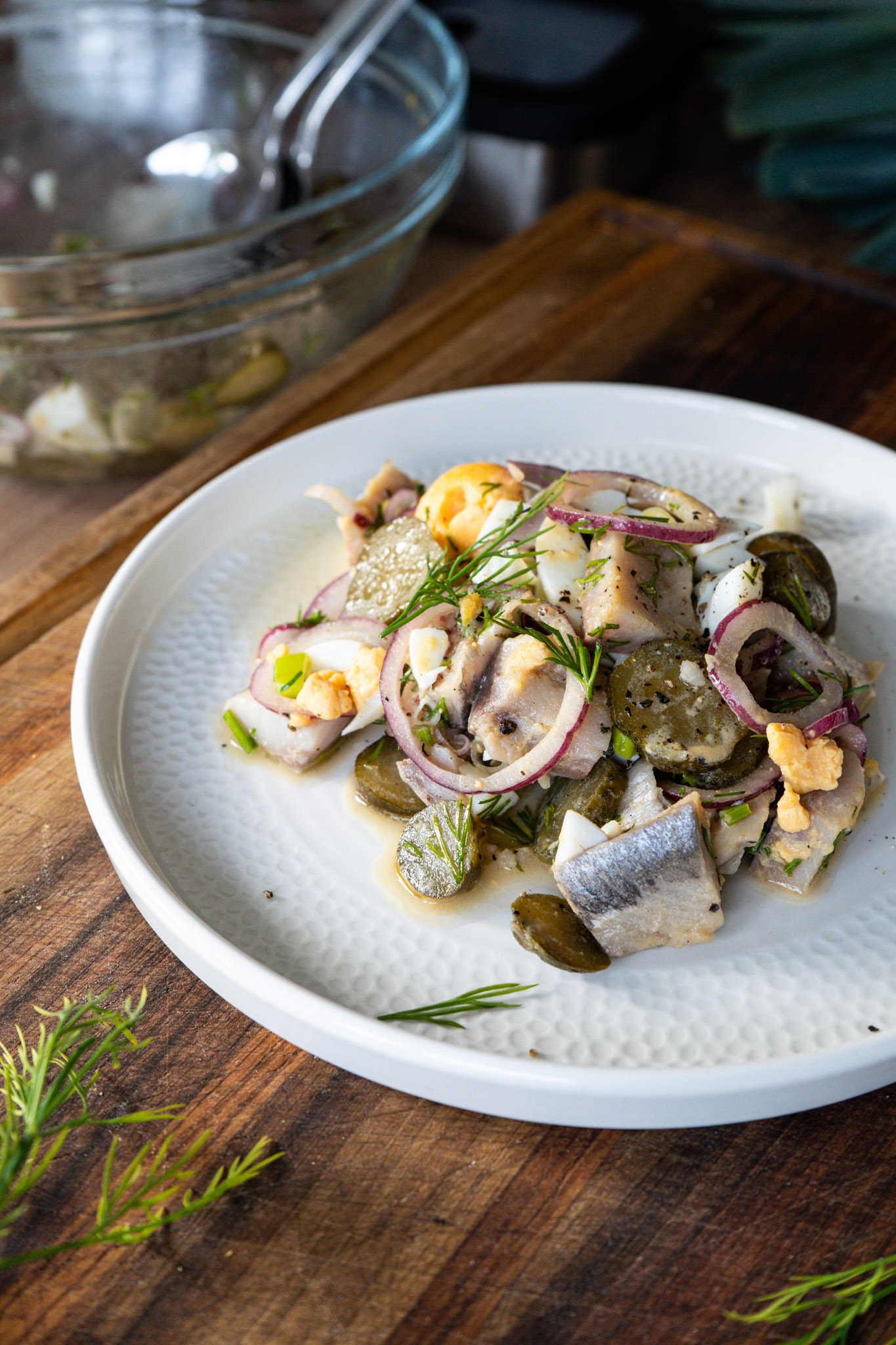 Heringstatar mit Gurken, Zwiebeln und Kräutern auf weißem Teller, angerichtet auf einem Holzbrett.