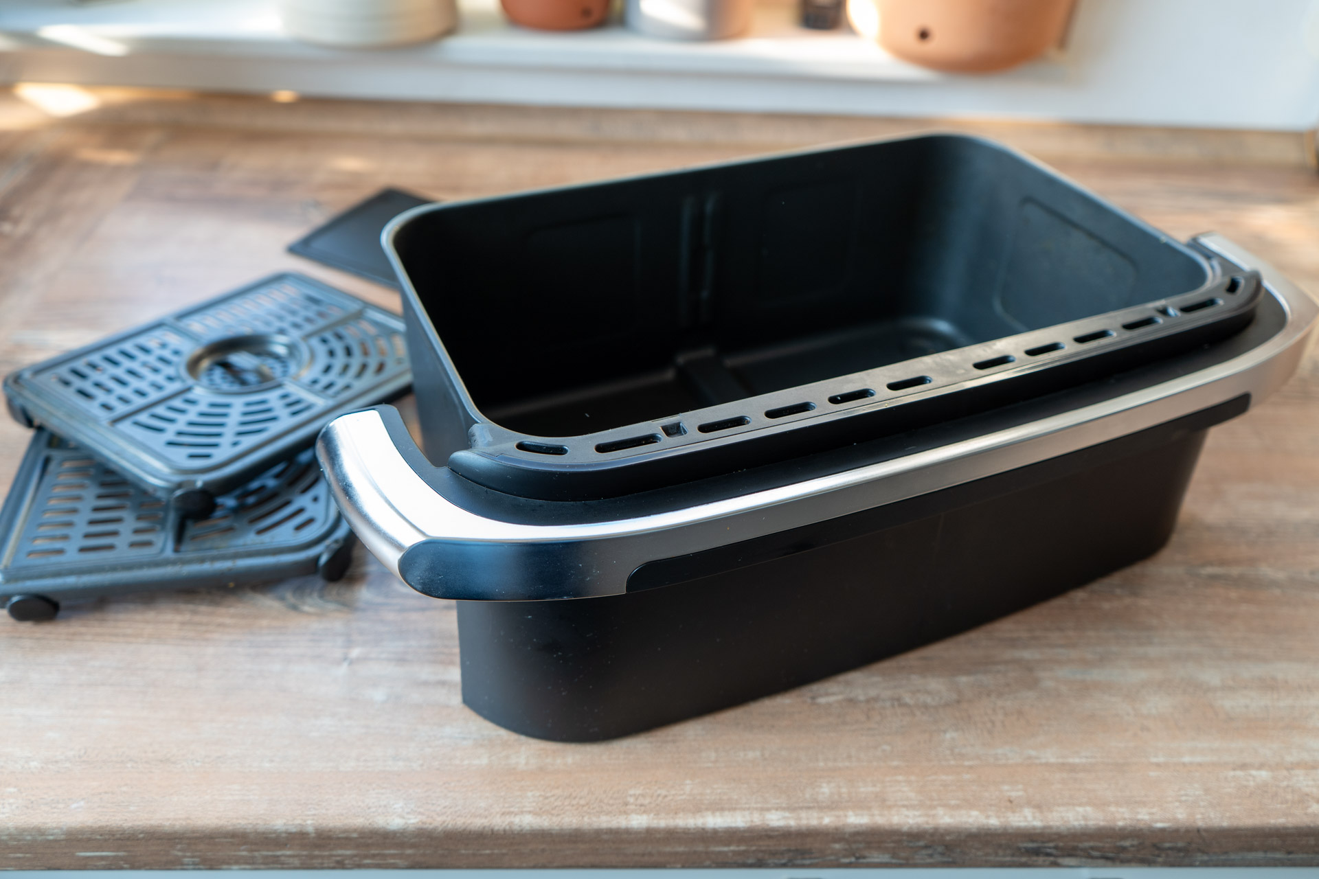 Ninja Foodi FlexDrawer AF500EU Test_05 Schwarzer Abtropfbehälter und Gittereinsätze auf einem Holztisch in einer Küche.