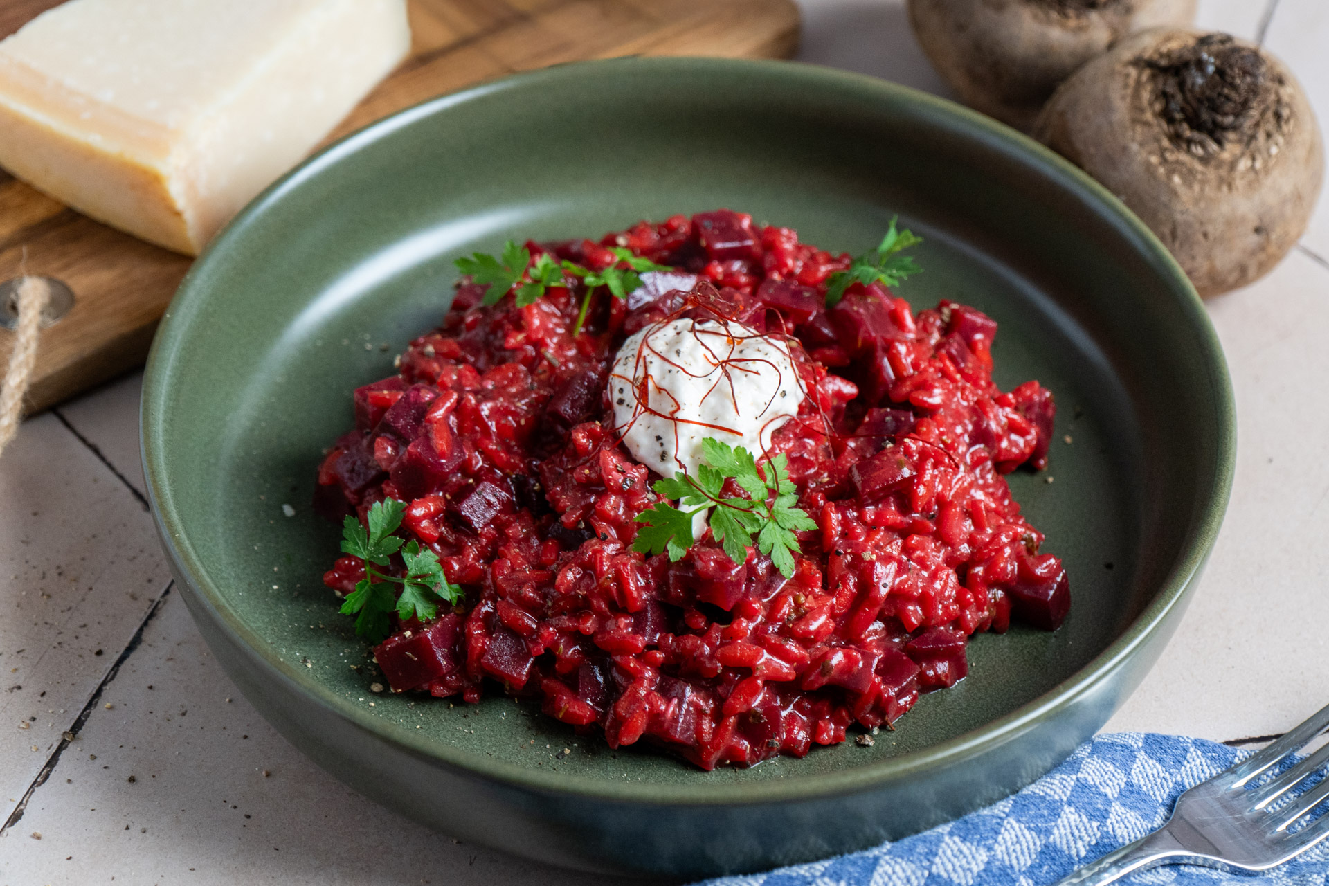 Rote_Bete_Risotto_01 Rote-Bete-Risotto mit Sahnehäubchen und Kräutern auf grünem Teller, neben Käse und frischen Rote-Bete-Knollen.