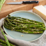 Grüner Spargel mit Käse überbacken auf einem Teller, daneben frische Spargelstangen, Käseblock und Messer auf Holzbrett.