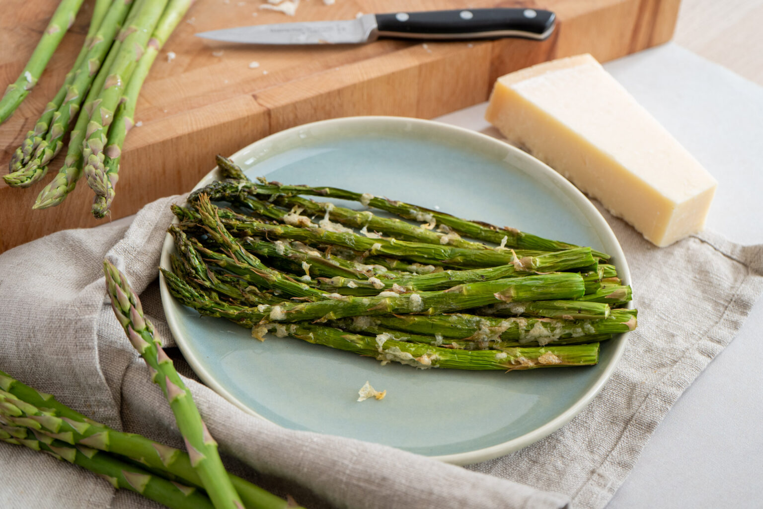 Grüner Spargel mit Käse überbacken auf einem Teller, daneben frische Spargelstangen, Käseblock und Messer auf Holzbrett.