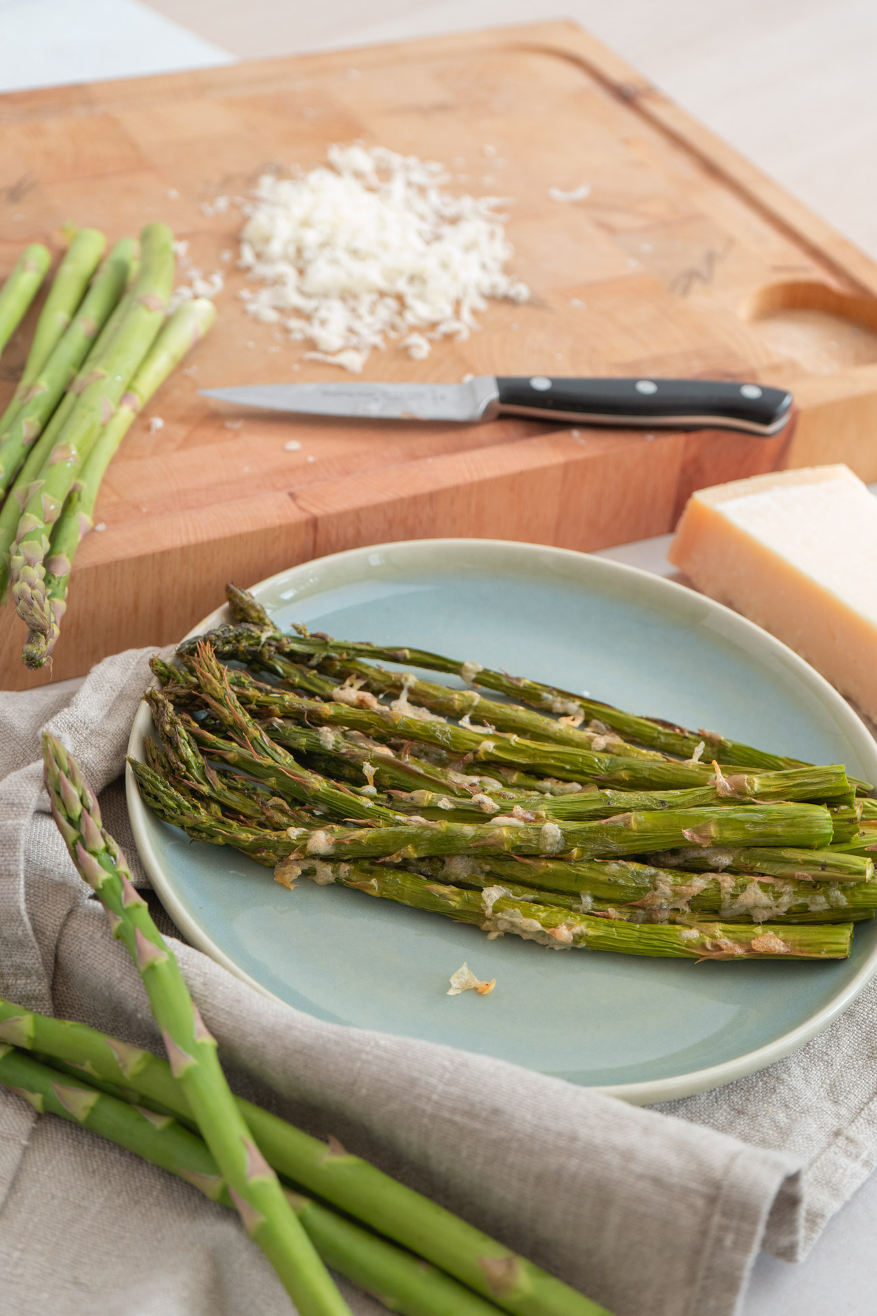 Gebackener grüner Spargel auf Teller, Holzbrett mit Messer, geriebener Käse und Käsestück im Hintergrund.