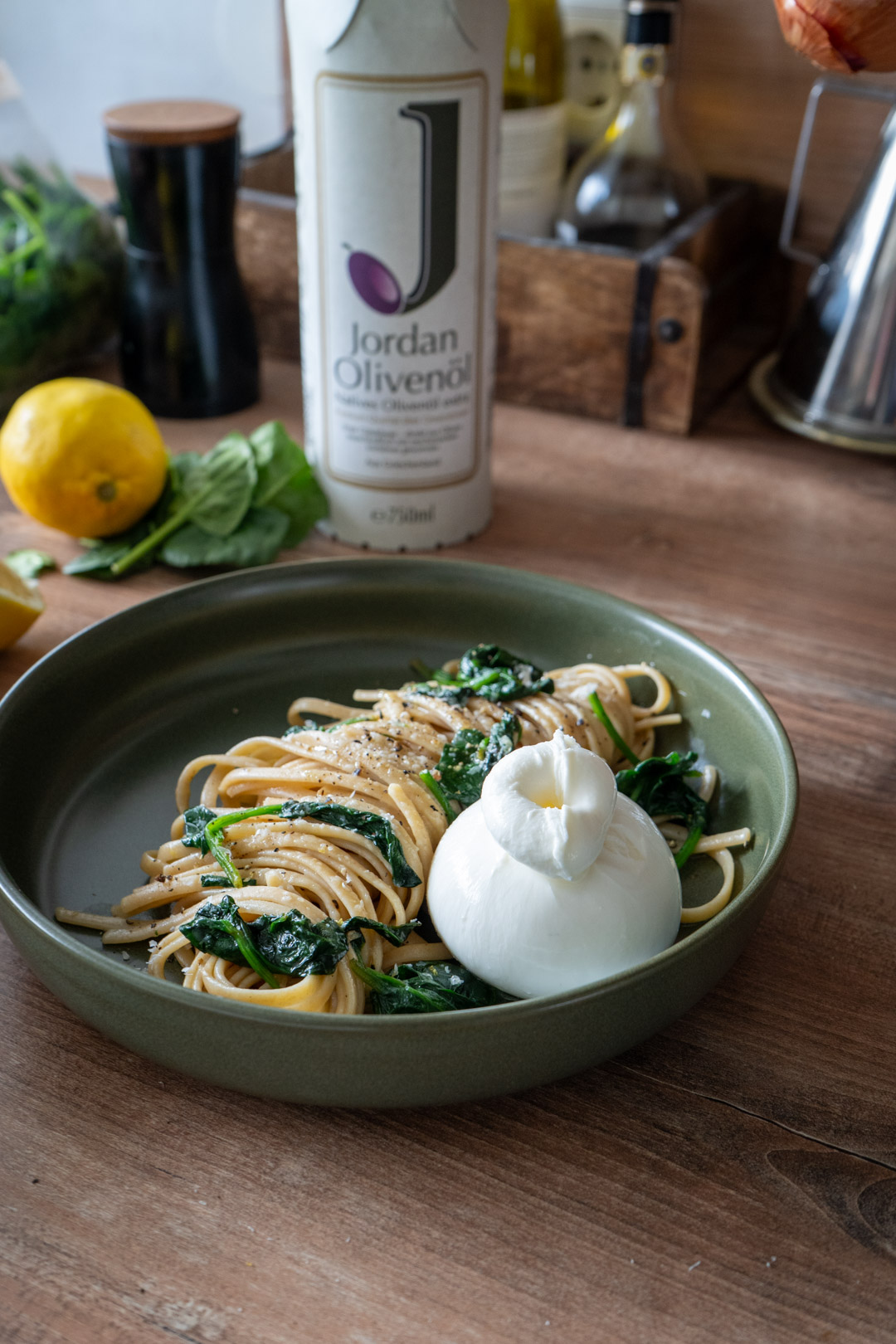 Pasta mit Spinat und Burrata in einer Schale, daneben Zitrone und Olivenöl auf einem hölzernen Tisch.