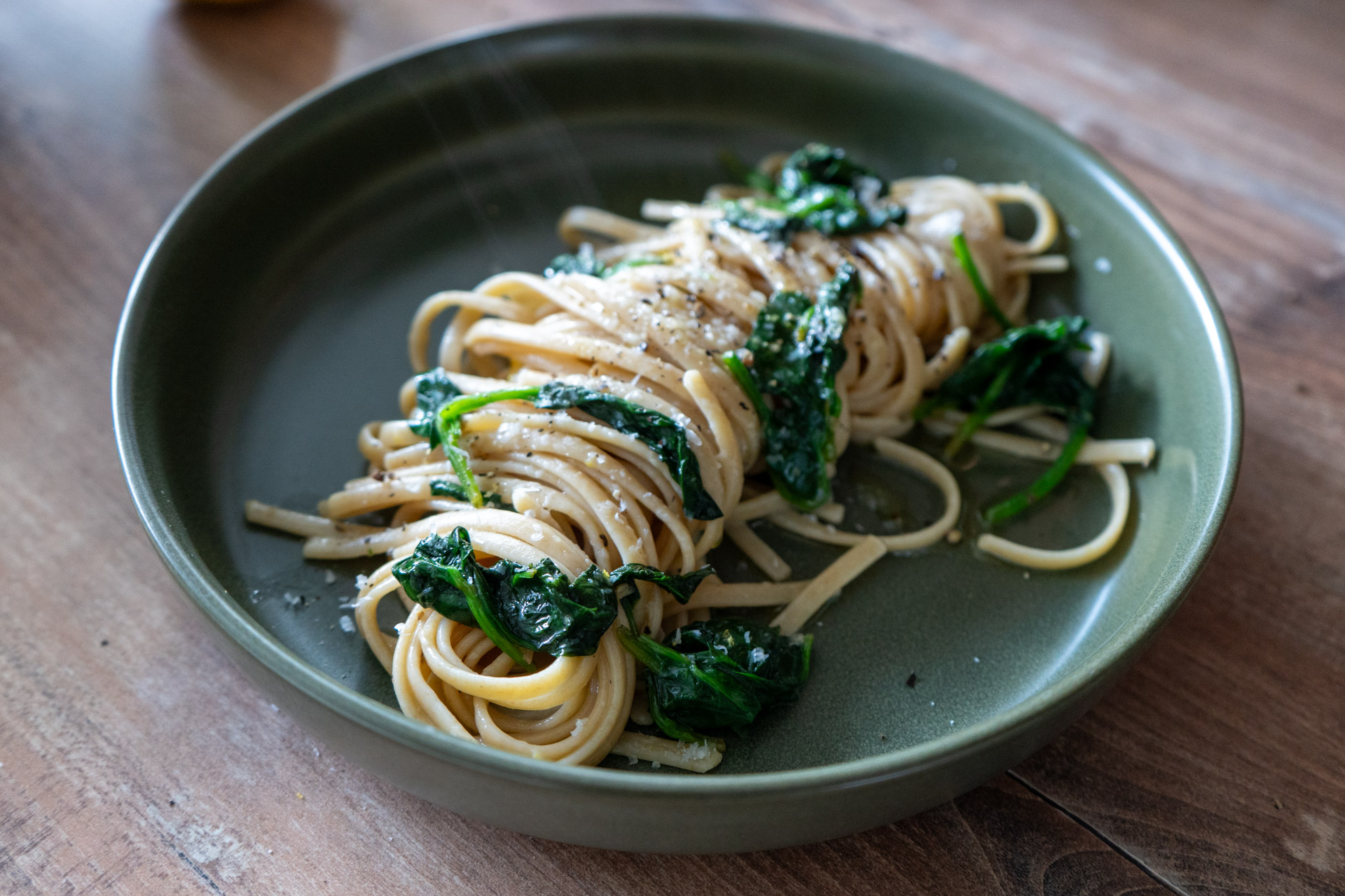 Nudeln mit Spinat in grüner Schale auf Holztisch, leicht mit Pfeffer bestreut.
