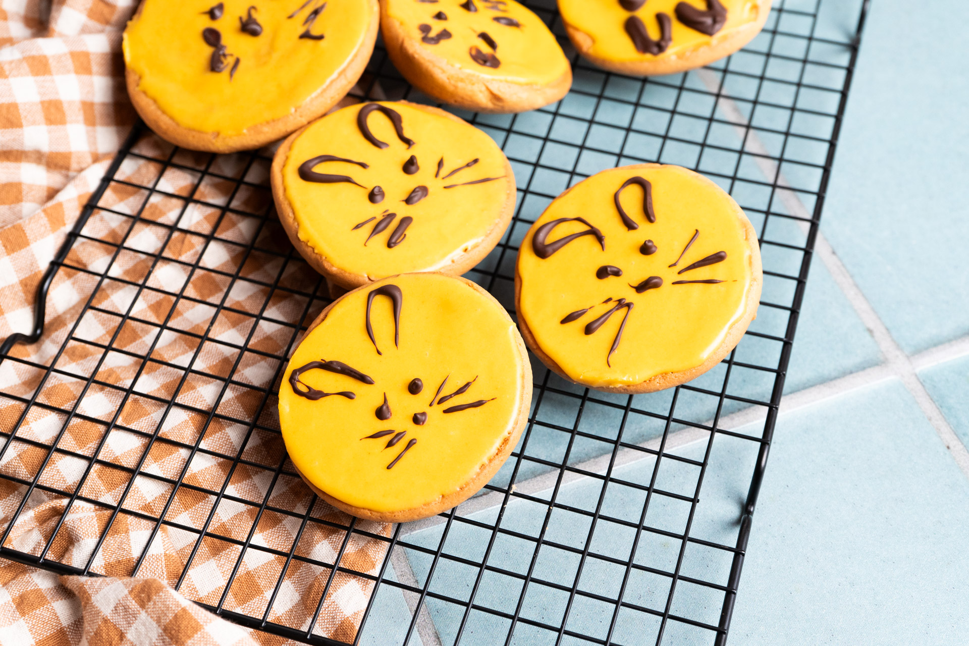 Kekse mit gelber Glasur auf einem Gitter. Mit Schokolade sind Hasengesichter verziert. Kariertes Tuch im Hintergrund.
