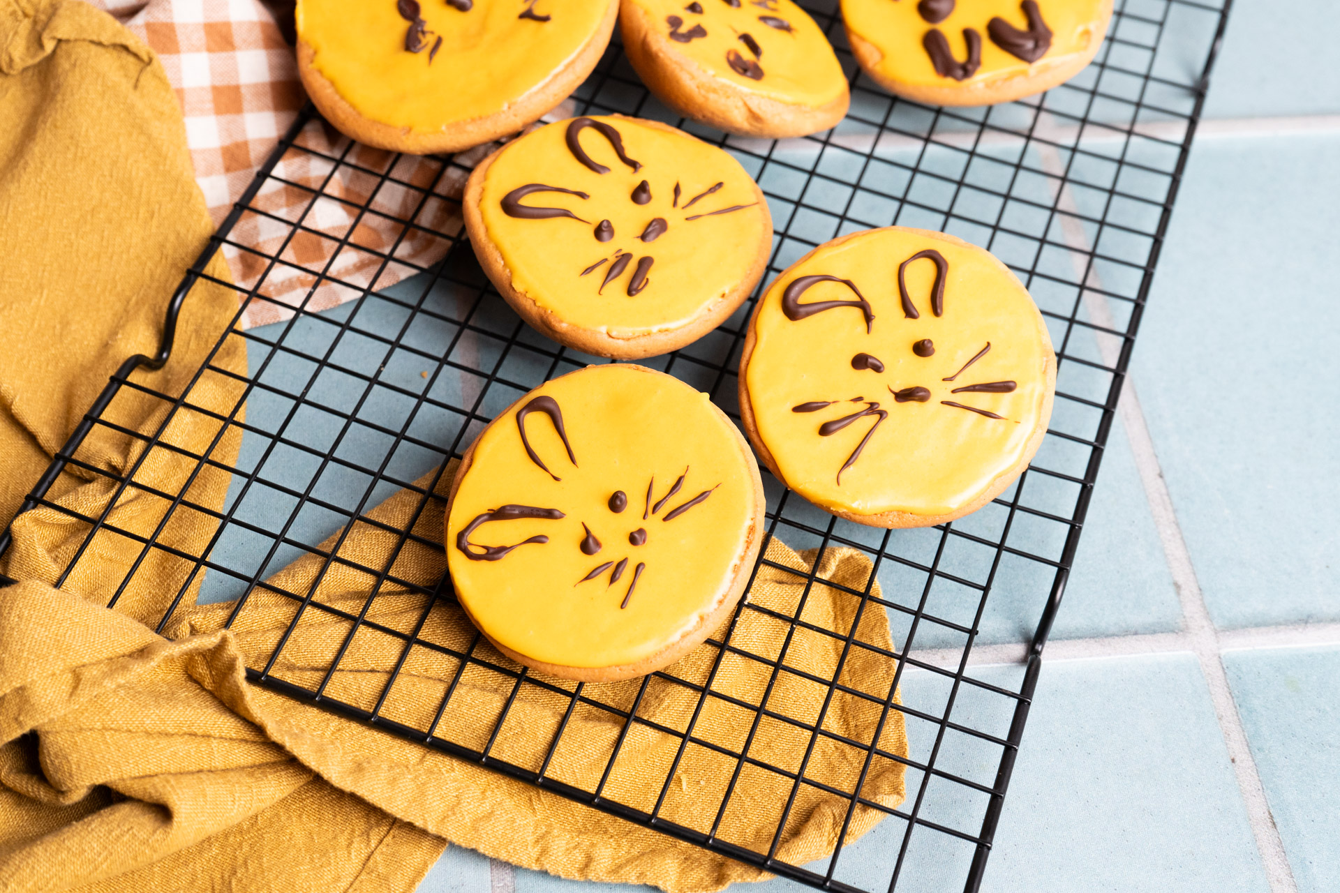 Gelbe Kekse mit Hasengesichtern aus Schokolade auf einem Gitter, daneben gelbes Tuch und karierter Stoff.