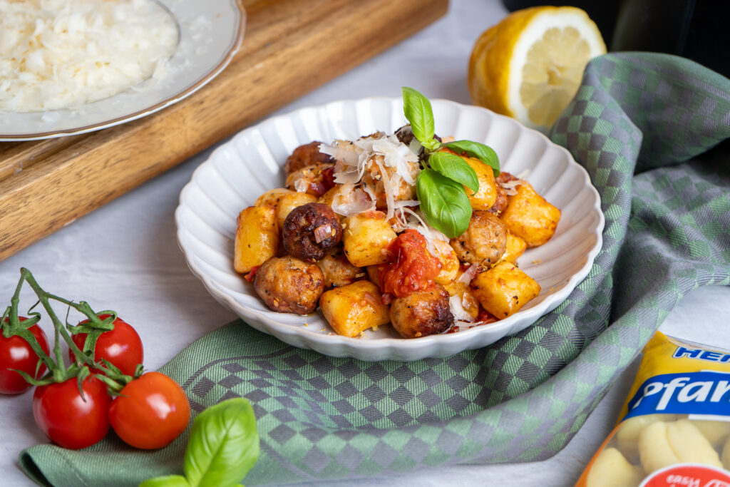 Weiße Schale mit Gnocchi, Fleischbällchen, Tomaten, Basilikum und Parmesan. Daneben Tomaten, Zitrone und ein Teller.