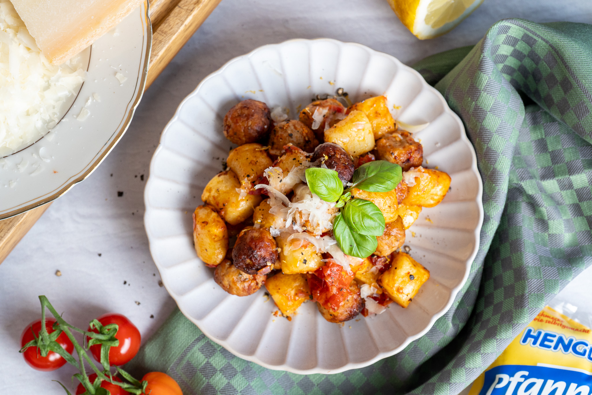 Pfannen-Gnocchi_03 Teller mit Gnocchi und Fleischbällchen, garniert mit Basilikum und Parmesan, daneben Tomaten und Parmesanblock.