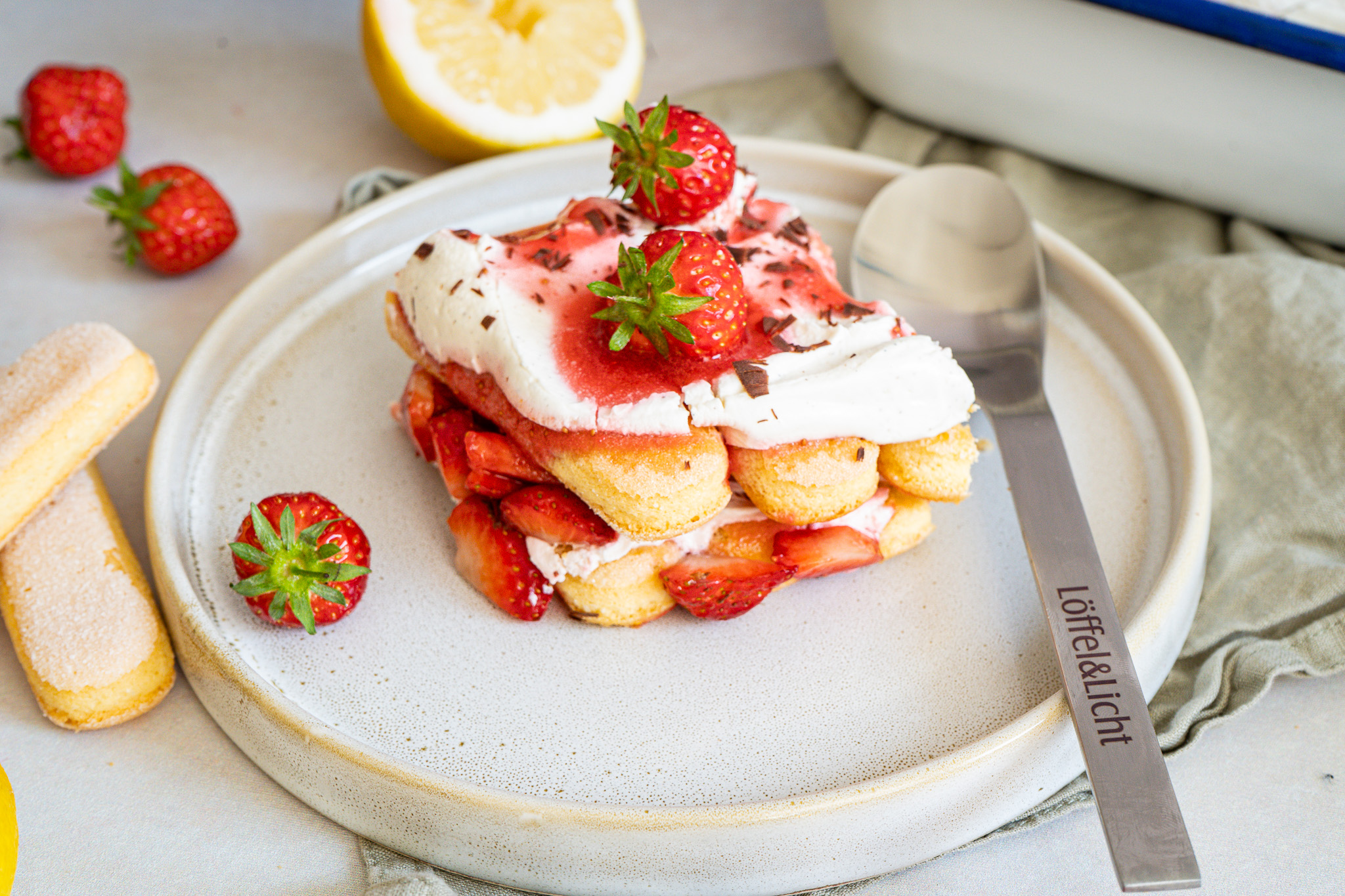 Dessert mit Löffelbiskuits, Sahne und frischen Erdbeeren auf einem Teller, dekoriert mit einem Löfel und halbierter Zitrone.