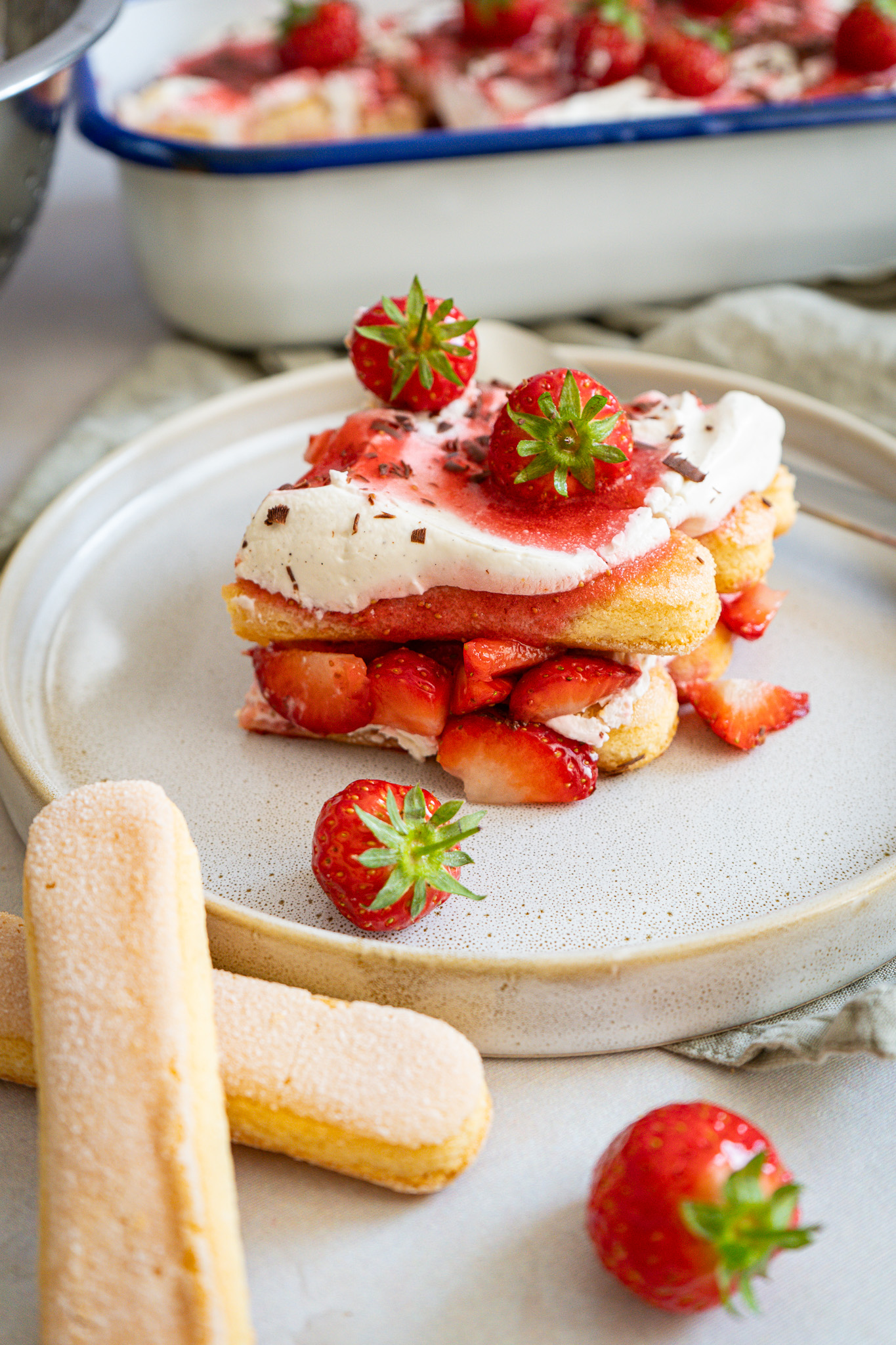 Erdbeer-Tiramisu mit Sahne auf Teller, garniert mit frischen Erdbeeren. Im Hintergrund weitere Portion in einer Schale.