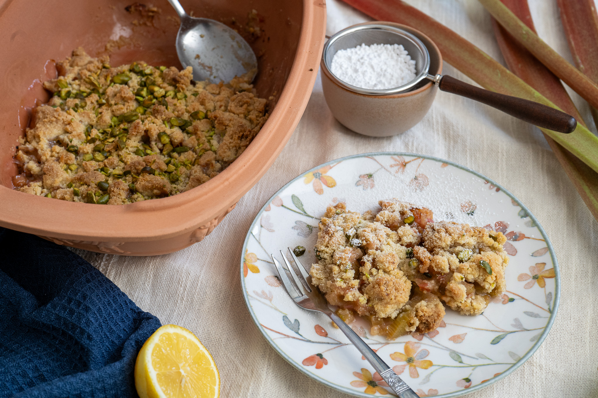 Keramikform mit Rhabarber-Crumble neben einem Teller mit Dessert, Puderzucker und halber Zitrone.