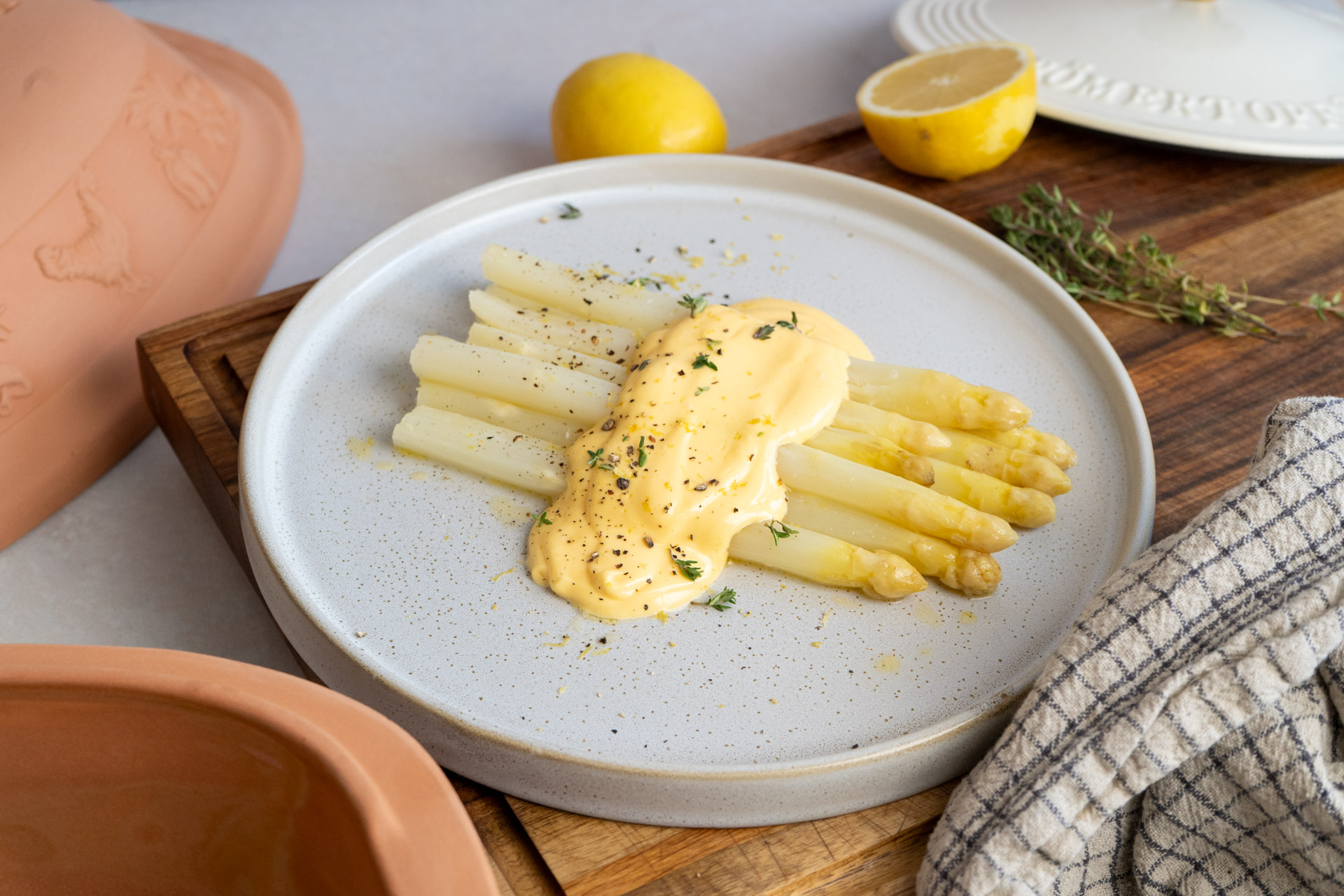 Weißer Spargel mit gelber Sauce auf einem Teller, garniert mit Kräutern, neben Zitrone auf Holzbrett platziert.