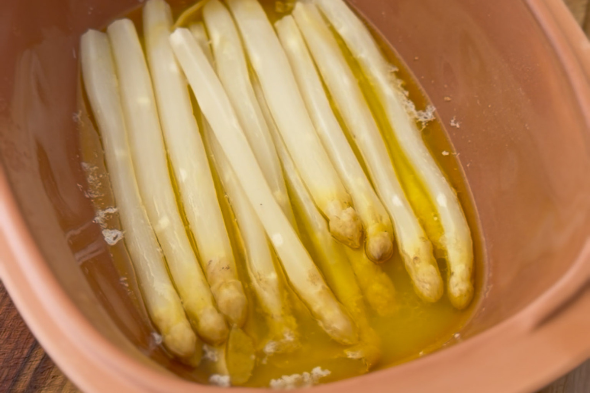Weiße Spargelstangen in einer braunen Auflaufform liegen in Flüssigkeit, möglicherweise Öl oder Brühe.