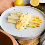 Weißer Spargel mit Sauce Hollandaise und frischen Kräutern, serviert auf einem weißen Teller, daneben halbe Zitrone.