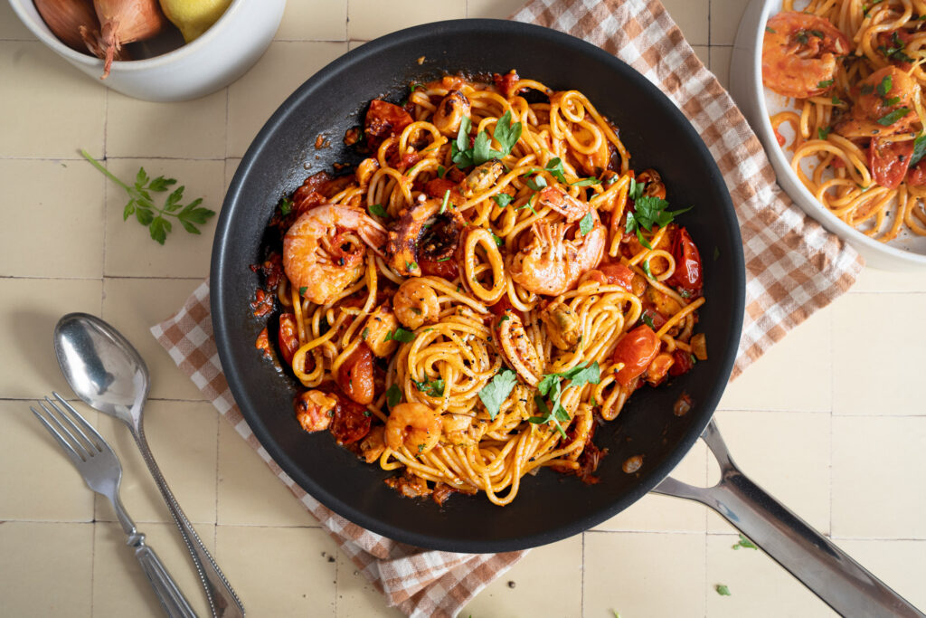 Pfanne mit Meeresfrüchte-Spaghetti in Tomatensoße, garniert mit Petersilie, auf kariertem Tuch, daneben Gabel und Löffel.