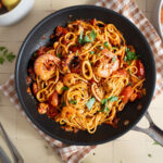 Pfanne mit Meeresfrüchte-Spaghetti in Tomatensoße, garniert mit Petersilie, auf kariertem Tuch, daneben Gabel und Löffel.