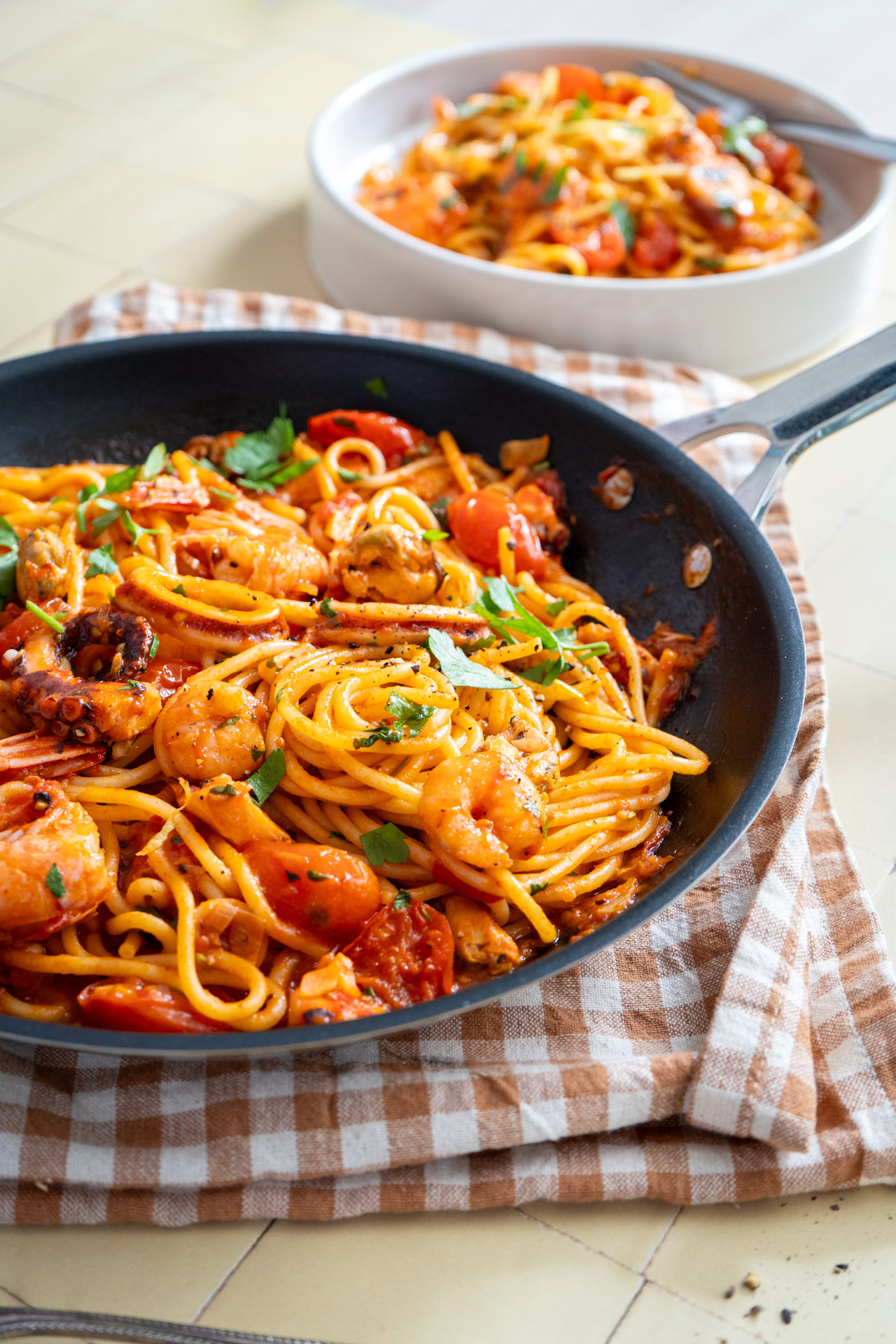 Bratpfanne mit Spaghetti und Meeresfrüchten in Tomatensoße, garniert mit Kräutern, auf einem karierten Tuch.