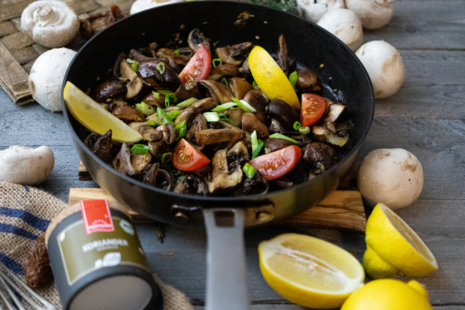 Antipasti-Champignons mit Cherrytomaten und Frühlingszwiebeln in einer Pfanne, daneben Zitronen und Champignons.