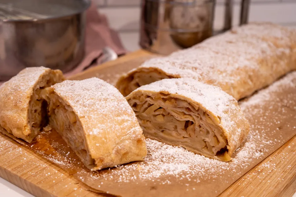 Angeschnittener Apfelstrudel mit Puderzucker auf Backpapier und Holzbrett, im Anschnitt die Apfelfüllung sichtbar.