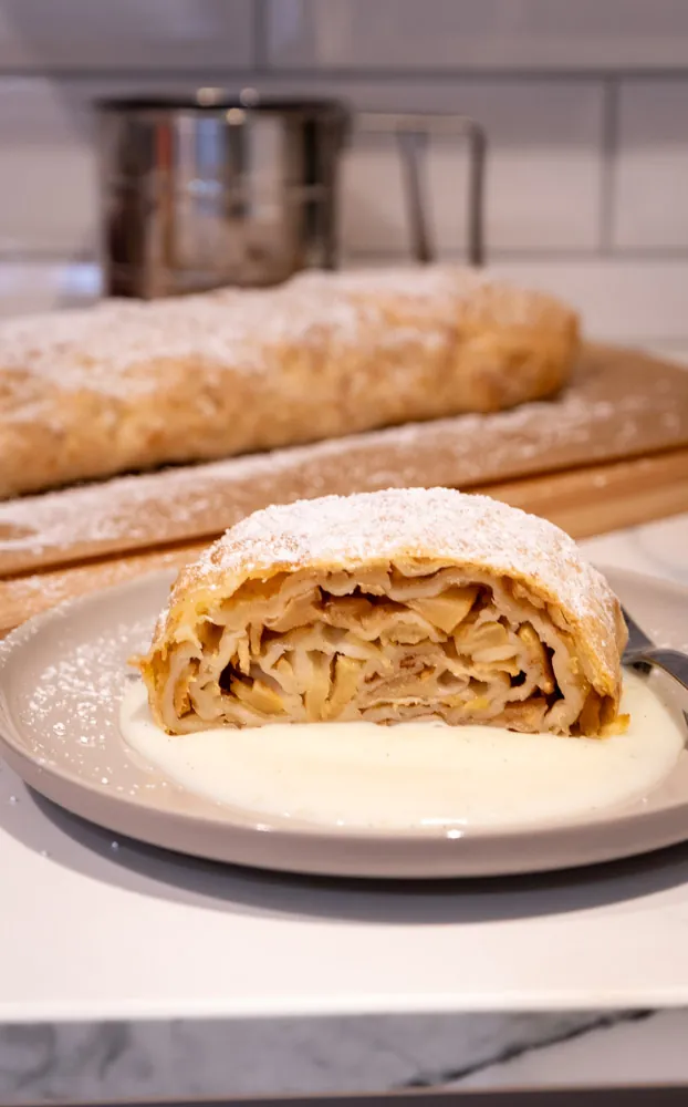 Ein Stück Apfelstrudel mit Vanillesauce auf einem grauen Teller im Hochformat, dahinter der Strudel.