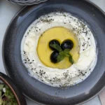 Labneh mit Olivenöl, schwarzen Oliven, Minze und Schwarzkümmel auf einem dunklen Teller, dahinter Pita im Korb.