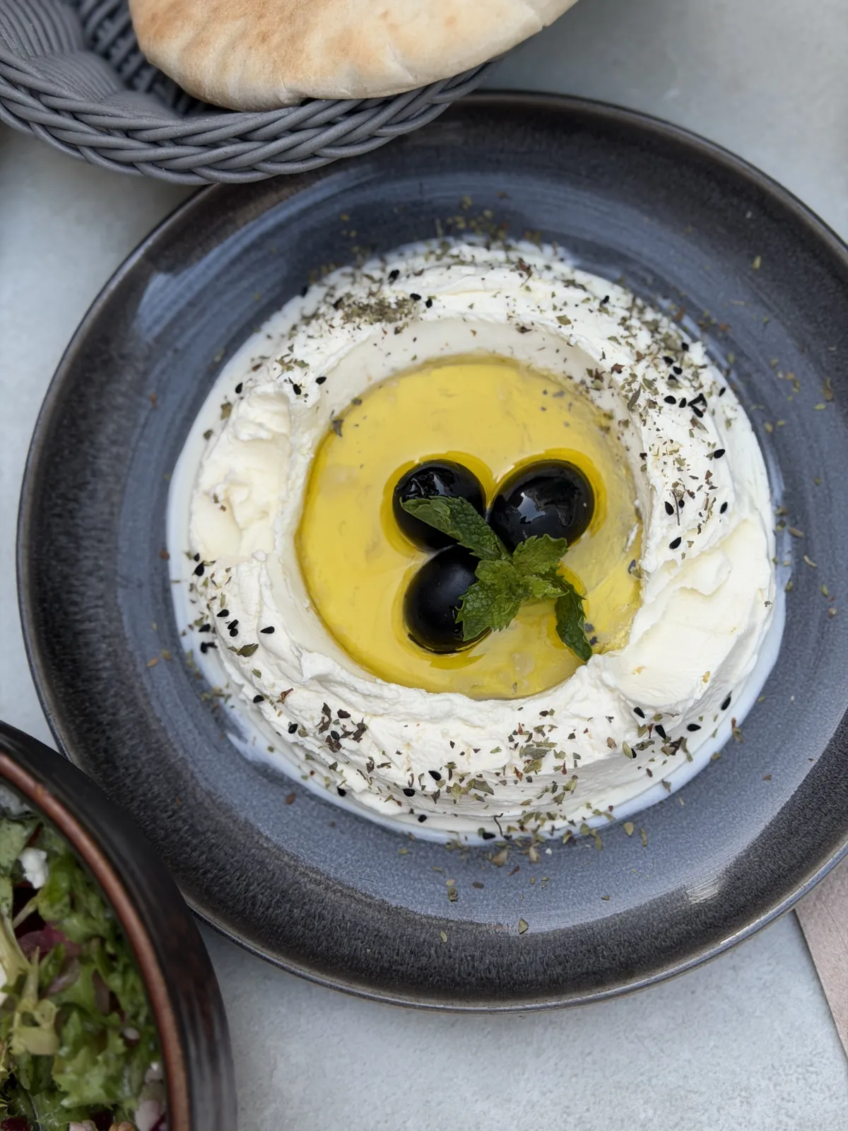 Labneh mit Olivenöl, schwarzen Oliven, Minze und Schwarzkümmel auf einem dunklen Teller, dahinter Pita im Korb.