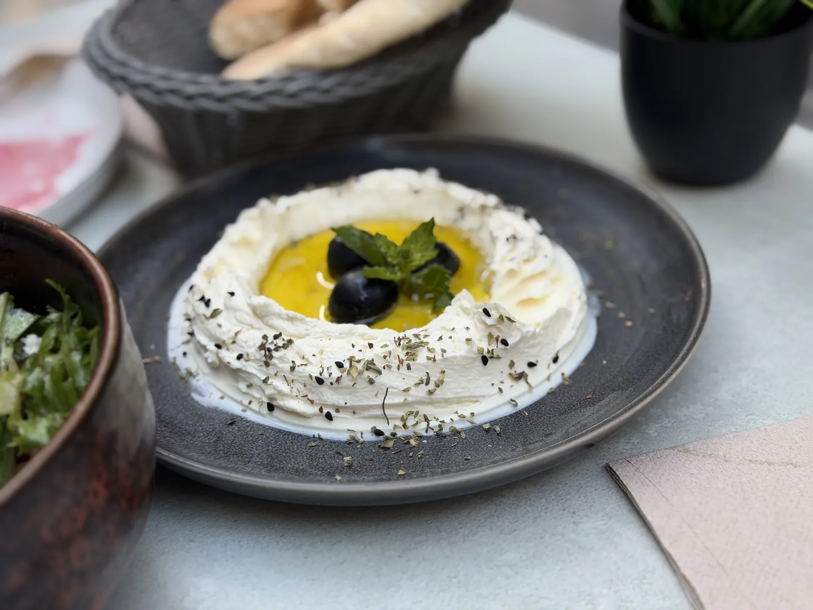 Labneh mit Olivenöl und Oliven auf einem Tisch, daneben Salat und ein Korb mit Pita.