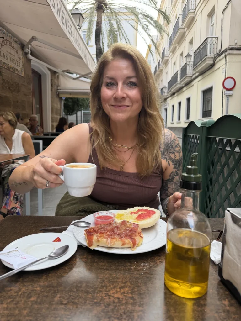 Christina sitzt lächelnd mit einer Kaffeetasse am Frühstückstisch in einer Gasse von Cádiz.