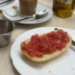 Geröstetes Brötchen mit geriebener Tomate auf einem weißen Teller, daneben ein Metallschälchen und Kaffee.