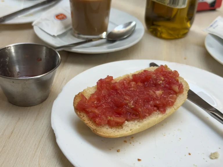 Geröstetes Brötchen mit geriebener Tomate auf einem weißen Teller, daneben ein Metallschälchen und Kaffee.