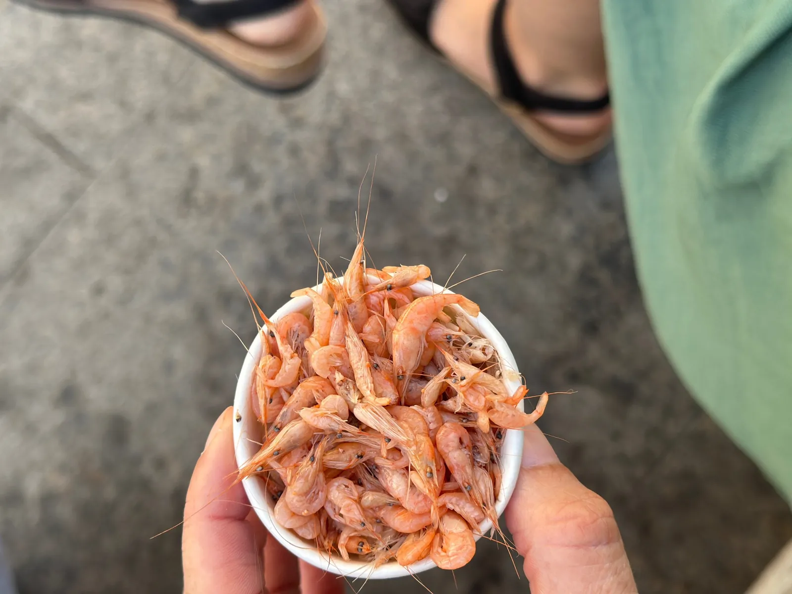Hand hält einen Becher mit kleinen Camarones über dem Boden der Markthalle.